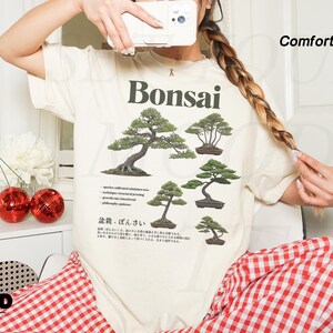 May include: Ivory-colored t-shirt with a Bonsai tree graphic. The shirt features multiple illustrations of bonsai trees and Japanese text. The words "Ivory" and "Comfort Colors" are printed on the top right of the shirt.