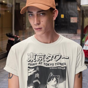 以下が含まれることがあります： 東京タワーと白黒の猫のグラフィックが描かれたベージュのTシャツ。Tシャツのテキストは「Paws at Tokyo Tower」と「Since 1958」です。その人は茶色の野球帽をかぶっています。
