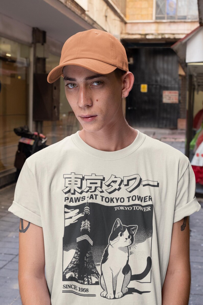 以下が含まれることがあります： 東京タワーと白黒の猫のグラフィックが描かれたベージュのTシャツ。Tシャツのテキストは「Paws at Tokyo Tower」と「Since 1958」です。その人は茶色の野球帽をかぶっています。