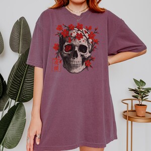 May include: A mauve-colored t-shirt featuring a detailed skull graphic adorned with red cherry blossoms. The skull has one red eye and is framed by the flowers. Japanese text is visible on the lower left.