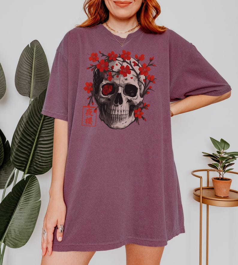 May include: A mauve-colored t-shirt featuring a detailed skull graphic adorned with red cherry blossoms. The skull has one red eye and is framed by the flowers. Japanese text is visible on the lower left.