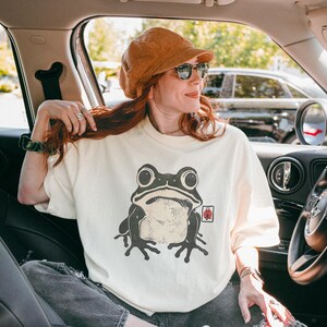 May include: Cream-colored t-shirt featuring a large, stylized frog graphic in shades of gray and black. The frog has big eyes and a detailed design. The shirt is worn by a person in a car.