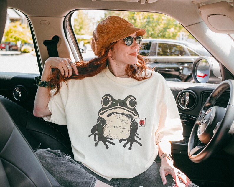 May include: Cream-colored t-shirt featuring a large, stylized frog graphic in shades of gray and black. The frog has big eyes and a detailed design. The shirt is worn by a person in a car.