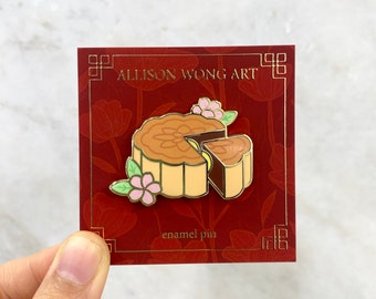 May include: An enamel pin featuring a mooncake with a slice removed, decorated with pink flowers and green leaves. The pin is attached to a dark red backing card with gold text that reads ALLISON WONG ART and enamel pin in a gold frame.