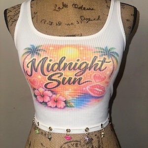 May include: White ribbed tank top with a colorful graphic that reads "Midnight Sun". The design includes palm trees, a sunset, flowers, and lips. A decorative chain with charms is attached at the waist.