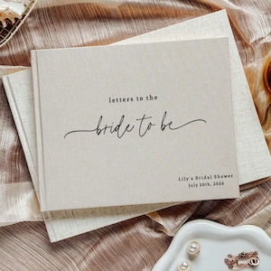 Puede incluir: Un libro beige cubierto de lino con el texto "letters to the bride to be" en escritura negra. El libro está abierto sobre una tela dorada. También se ven una pequeña vela en un portavelas y una bandeja blanca con joyas.