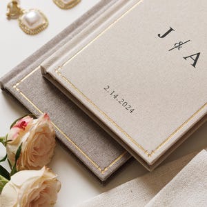 May include: Two rectangular books with beige and light brown fabric covers, featuring gold-stitched borders. The top book displays the black text J & A and the date 2.14.2024. Two square pearl earrings with gold frames and two pale pink roses are nearby.