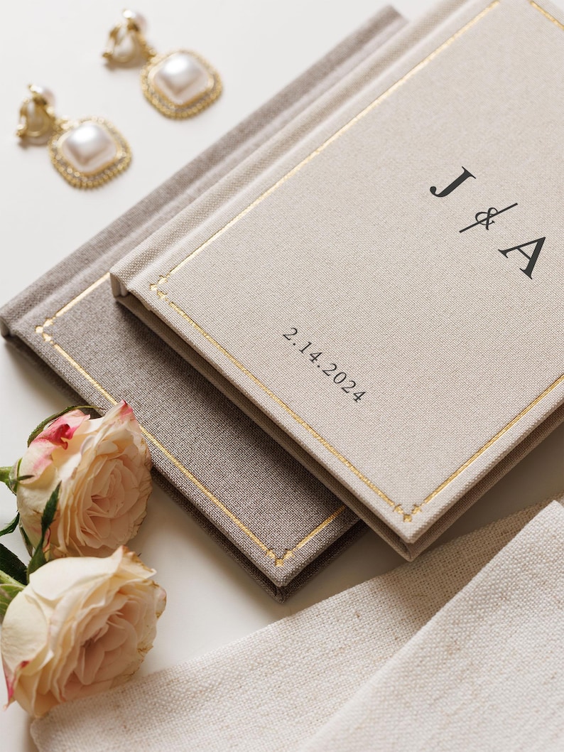 May include: Two rectangular books with beige and light brown fabric covers, featuring gold-stitched borders. The top book displays the black text J & A and the date 2.14.2024. Two square pearl earrings with gold frames and two pale pink roses are nearby.