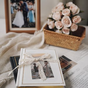 May include: A white hardback photo album with gold-toned edges, tied with a cream-coloured lace ribbon. The cover features a rectangular photograph of a couple on a beach. The album rests on a black book, accompanied by a bouquet of pale pink roses.