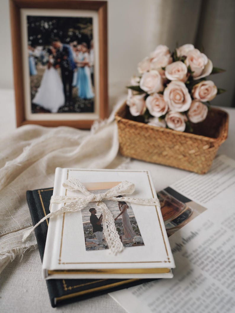 May include: A white hardback photo album with gold-toned edges, tied with a cream-coloured lace ribbon. The cover features a rectangular photograph of a couple on a beach. The album rests on a black book, accompanied by a bouquet of pale pink roses.