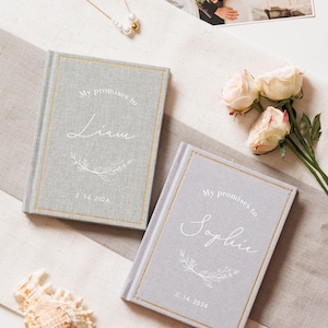May include: Two light grey fabric-covered vow books with gold-foiled borders. The covers feature the white printed text "My promises to" above the names "Liam" and "Sophie" respectively, with a floral branch graphic and the date "2. 14. 2024".