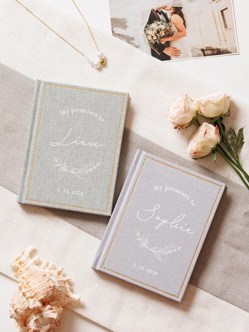 May include: Two light grey fabric-covered vow books with gold-foiled borders. The covers feature the white printed text "My promises to" above the names "Liam" and "Sophie" respectively, with a floral branch graphic and the date "2. 14. 2024".