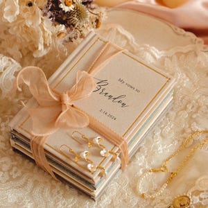 May include: A stack of two small hardcover books with beige covers, tied together with a peach-coloured organza ribbon. The top book features the text My vows to Brandon and the date 2.14.2024. Gold-toned earrings with small white pearls rest on top.