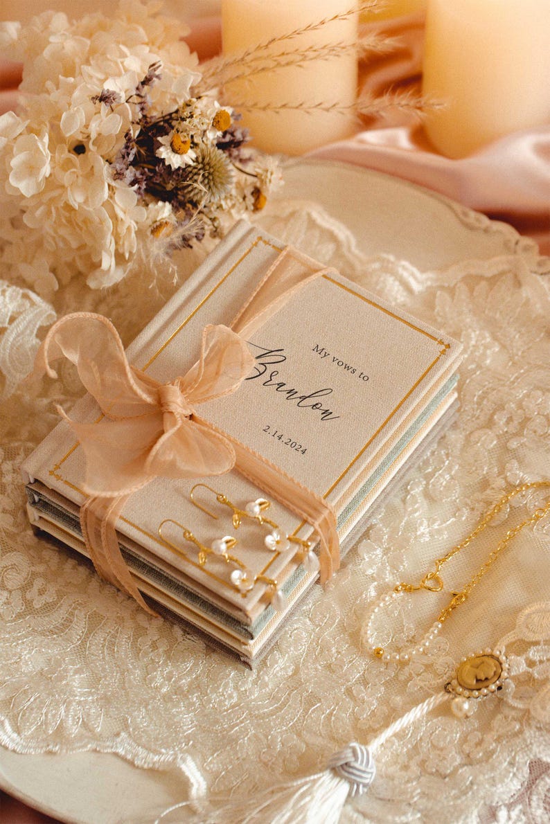 May include: A stack of two small hardcover books with beige covers, tied together with a peach-coloured organza ribbon. The top book features the text My vows to Brandon and the date 2.14.2024. Gold-toned earrings with small white pearls rest on top.