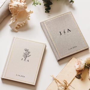 May include: Two rectangular hardcover books with beige fabric covers, gold foil border detailing, and black printed text. One book features the initials J & A with the date 2.14.2024, while the other displays a floral illustration, J A, and 2.14.2024.