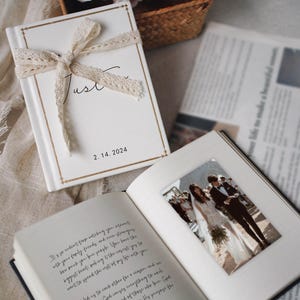 May include: A white hardback book with a cream lace ribbon tied in a bow, featuring gold border lines and the text "Just" and "2. 14. 2024". The open book displays a printed photograph of a couple and handwritten text on the opposite page.