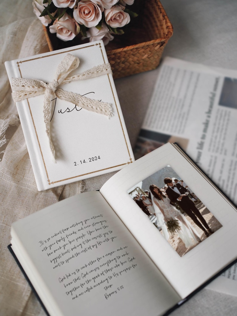 May include: A white hardback book with a cream lace ribbon tied in a bow, featuring gold border lines and the text "Just" and "2. 14. 2024". The open book displays a printed photograph of a couple and handwritten text on the opposite page.