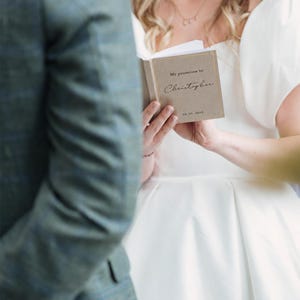 May include: A small, rectangular, tan-coloured vow book with gold-foiled text that reads My promises to Christopher and the date 04.20.2024. The book is held open, displaying a clean white page on the left side and the printed cover on the right.