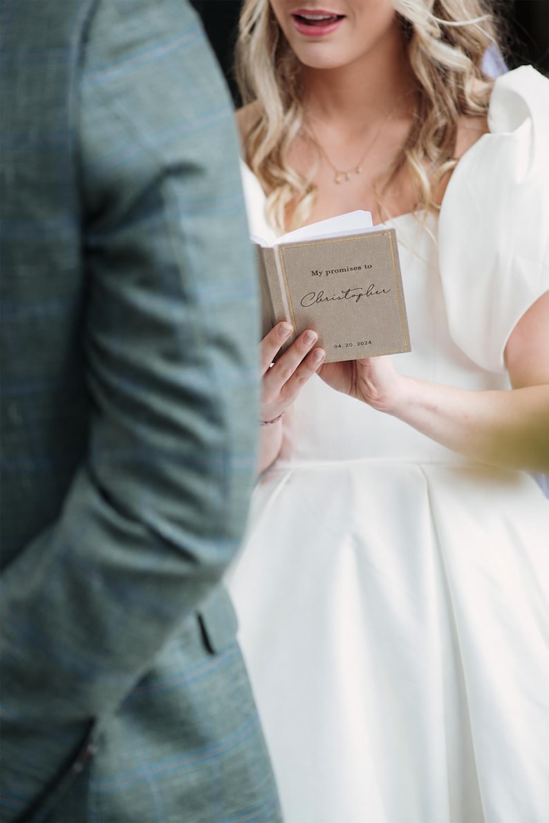 May include: A small, rectangular, tan-coloured vow book with gold-foiled text that reads My promises to Christopher and the date 04.20.2024. The book is held open, displaying a clean white page on the left side and the printed cover on the right.