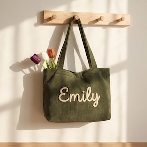May include: Olive green corduroy tote bag with the name "Emily" embroidered in a cream-colored script. The bag is hanging on a wooden coat rack and contains three tulips in shades of purple, white, and orange.