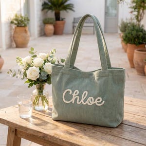 May include: Sage green corduroy tote bag with the name "Chloe" embroidered in white script. The bag has two handles and is sitting on a wooden table. A bouquet of white roses and a glass of water are next to the bag.
