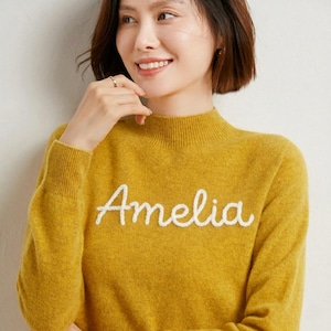 May include: Mustard yellow sweater with the name "Amelia" embroidered in white script across the chest. The sweater has a mock neck and long sleeves. The person in the image is smiling.