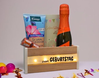 May include: A wooden gift box with a bottle of Prosecco, a tube of lotion, and a card. The box has the words "zum GEBURTSTAG" illuminated on the front. Colorful decorative eggs and confetti are scattered around the box.