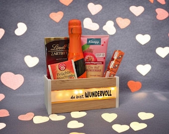 May include: A gift basket with a bottle of Prosecco, chocolate, tea, and body wash. The wooden box has the words "du bist WUNDERVOLL" illuminated on the front. Heart-shaped confetti decorates the background.