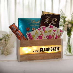 Op de afbeelding: Een houten geschenkdoos gevuld met chocolaatjes, theezakjes en een chocoladereep. De doos heeft een lichtstrip met de tekst "eine KLEINIGKEIT". Een boeket witte bloemen staat op de achtergrond.