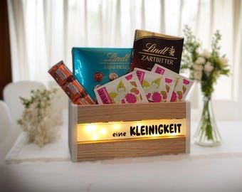 May include: A wooden gift box filled with chocolates, tea packets, and a chocolate bar. The box has a light strip with the text "eine KLEINIGKEIT". A bouquet of white flowers is in the background.