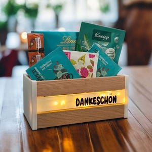 May include: A wooden box with a white frame and a light-up panel displaying the word "DANKESCHÖN". The box contains tea bags, chocolates, and other small items. The box is on a wooden table.
