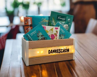 May include: A wooden box with a white frame and a light-up panel displaying the word "DANKESCHÖN". The box contains tea bags, chocolates, and other small items. The box is on a wooden table.