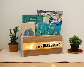 May include: A wooden box with the words "gute BESSERUNG" illuminated on the front. The box contains tea packets, a small plant, and other items. The box is on a white surface.