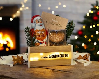 May include: A wooden box with the words "frohe WEIHNACHTEN" illuminated on the front. Inside, a Santa Claus chocolate figure and a packaged cookie are visible. The background includes a Christmas tree and a fireplace.