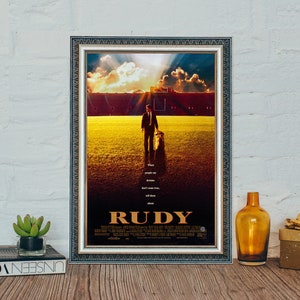 May include: A framed movie poster for the film "Rudy". The poster features a man in a suit walking across a football field with a dog. The poster has a vintage look with a faded yellow and orange color scheme. The text on the poster reads "When people say dreams don't come true, tell them about Rudy."