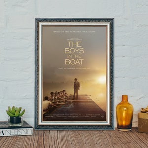 May include: A movie poster for the film "The Boys in the Boat" based on a true story. The poster features a group of men in a rowing boat on a lake at sunset. The title of the film is in large, bold letters at the top of the poster. The poster also includes the names of the director, George Clooney, and the actors, Alexandre Desplat, Grant Heslov, and Mark L. Smith.