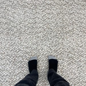 Puede incluir: Una alfombra texturizada de color blanco roto con una superficie densa y nudosa. El patrón de la alfombra está formado por pequeñas formas redondeadas. Se ven calcetines negros con punteras grises y pantalones negros.
