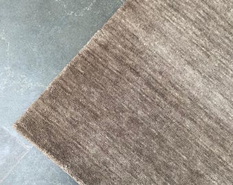 May include: A corner of a brown rug with a textured, striped pattern. The rug is a neutral color and appears to be made of a soft material. The rug is placed on a gray tile floor.