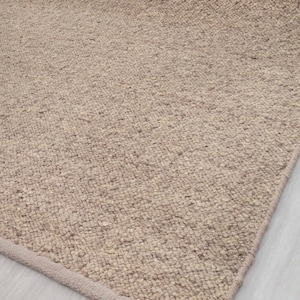 Puede incluir: Una alfombra beige con una superficie texturizada y tejida. El borde de la alfombra es visible, mostrando un borde definido. La vista de cerca enfatiza la textura tejida, ideal para la decoración de interiores.