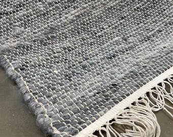 May include: Close-up of a handwoven rug with a textured surface. The rug features a mix of grey and white threads, creating a speckled pattern. The edge has a white border with long, off-white fringe.