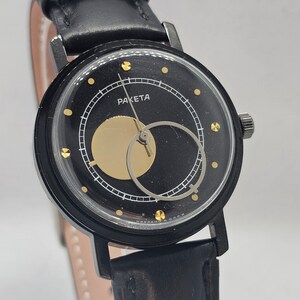 Puede incluir: Un reloj de pulsera negro con una correa de cuero negro. La esfera del reloj es negra con detalles dorados y la palabra "RAKETA" impresa en ella.