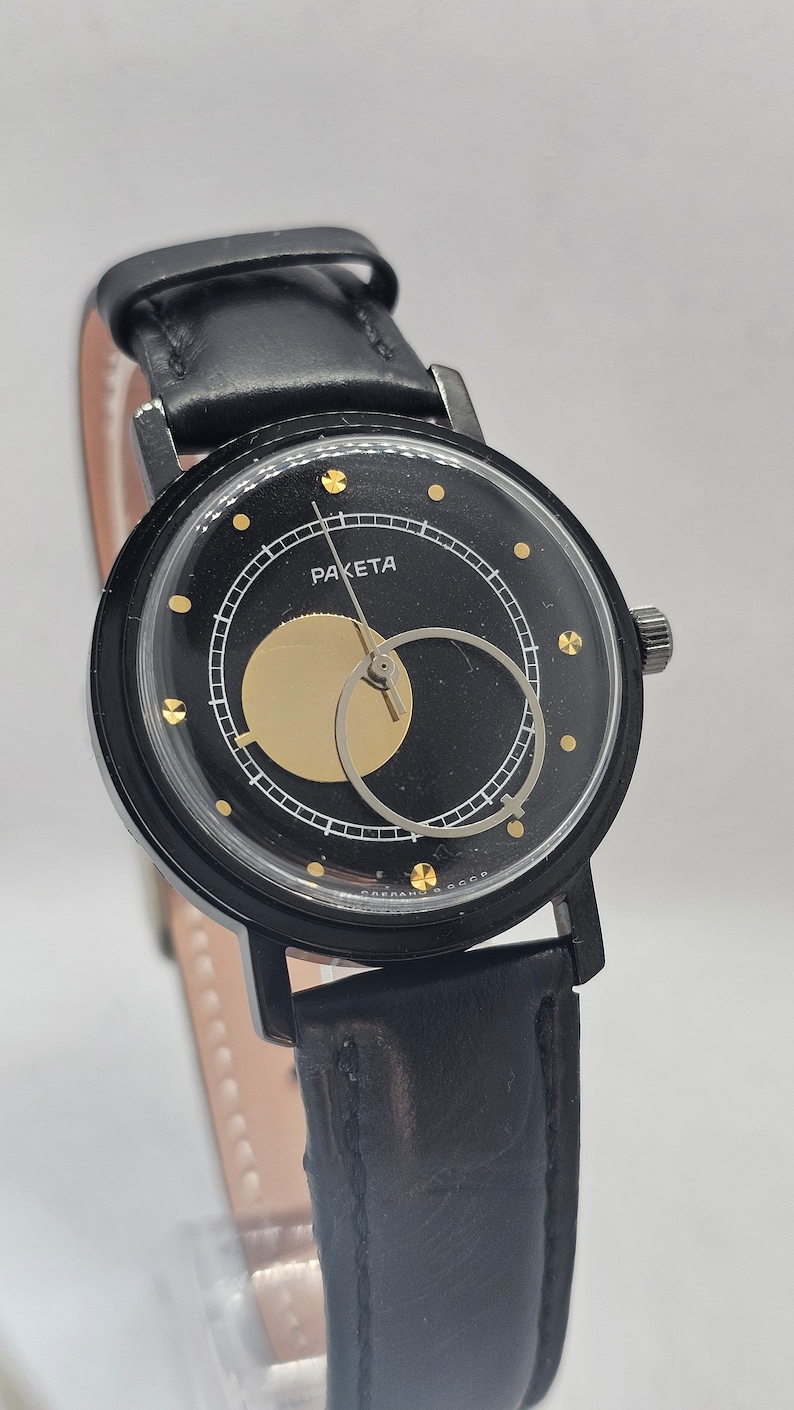Puede incluir: Un reloj de pulsera negro con una correa de cuero negro. La esfera del reloj es negra con detalles dorados y la palabra "RAKETA" impresa en ella.