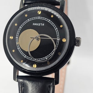 Puede incluir: Un reloj de pulsera negro con una correa de cuero negro. La esfera del reloj es negra con detalles dorados y presenta un dise&ntilde;o &uacute;nico con un c&iacute;rculo dorado que cubre parcialmente las manecillas del reloj. La esfera del reloj tambi&eacute;n tiene la palabra "PAKEТА" impresa en ella.