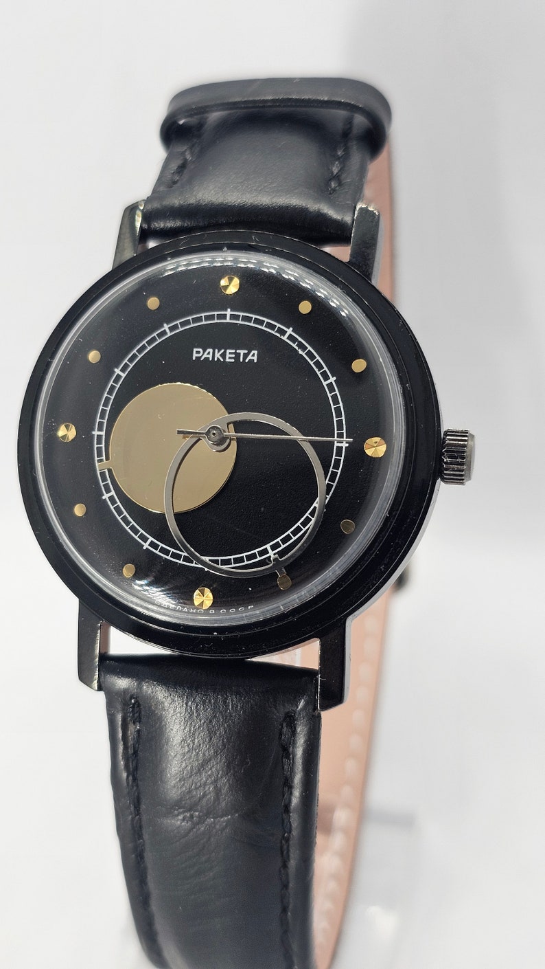 Puede incluir: Un reloj de pulsera negro con una correa de cuero negro. La esfera del reloj es negra con detalles dorados y presenta un dise&ntilde;o &uacute;nico con un c&iacute;rculo dorado que cubre parcialmente las manecillas del reloj. La esfera del reloj tambi&eacute;n tiene la palabra "PAKEТА" impresa en ella.