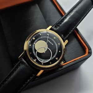 May include: A gold-tone wristwatch with a black face and a black leather strap. The watch face has a white circle with a smaller black circle inside. The word "PAKETA" is printed on the watch face.