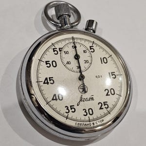 May include: A silver pocket watch with a white face and black hands. The watch has a black second hand and a small dial for measuring tenths of a second. The watch face has the words "СДЕЛАНО В СССР" and "Jkam" printed on it.