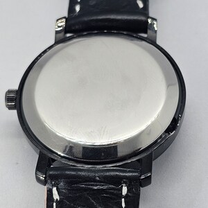 Puede incluir: La parte posterior de un reloj de pulsera negro con una caja plateada y una correa de cuero negro con costuras blancas. La correa tiene el n&uacute;mero "18" impreso en ella.