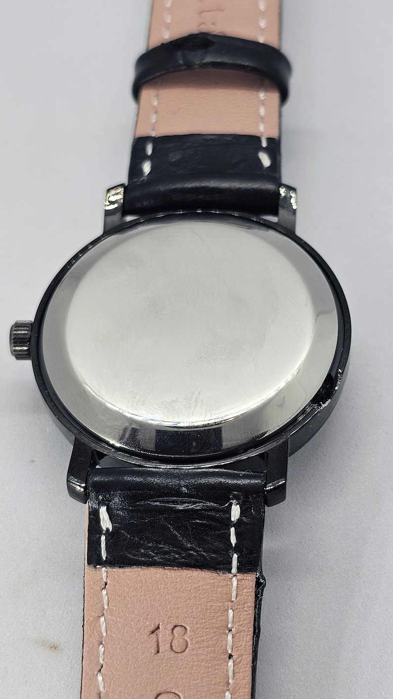 Puede incluir: La parte posterior de un reloj de pulsera negro con una caja plateada y una correa de cuero negro con costuras blancas. La correa tiene el n&uacute;mero "18" impreso en ella.