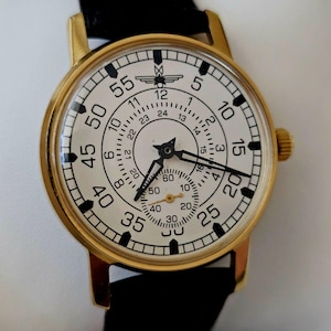Op de afbeelding: Een gouden polshorloge met een witte wijzerplaat en zwarte markeringen. Het horloge heeft een 24-uurs wijzerplaat en een kleinere wijzerplaat voor seconden. De wijzerplaat van het horloge heeft ook een klein vliegtuiglogo.
