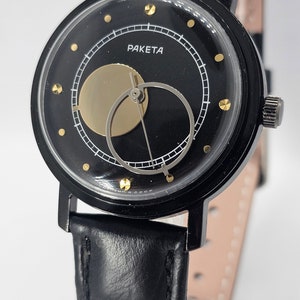 Puede incluir: Un reloj de pulsera negro con una correa de cuero negro. La esfera del reloj es negra con detalles dorados y presenta un dise&ntilde;o &uacute;nico de fase lunar. La esfera del reloj tambi&eacute;n tiene la palabra "PAKETA" impresa en ella.